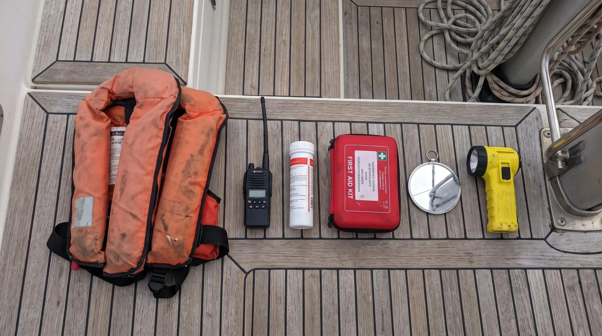 Vue en plongée sur pont teck de voilier avec équipements sécurité réglementaires disposés : gilets de sauvetage orange, VHF portable noire, conteneur fusées de détresse blanc, trousse premiers secours rouge, miroir signalisation, lampe torche étanche jaune, cordages en arrière-plan